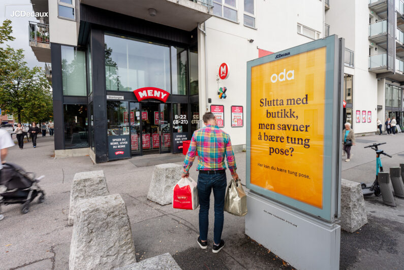 Person løfter bæreposer foran utendørsreklame for Oda.